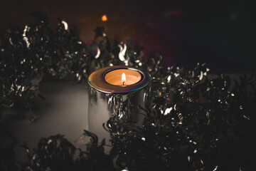 Burning candle in metal candle holder amongst silver tinsel for Christmas tree on dark background....