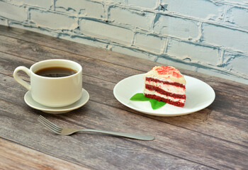 A plate with a piece of Red velvet cake decorated with mint leaves and a cup of hot black coffee on a saucer, the whole composition is on a light wooden table.