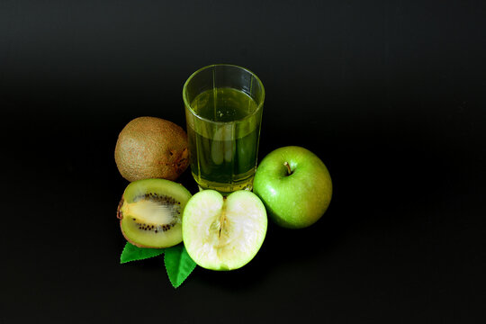 A tall glass of freshly squeezed fruit juice on a black background, next to ripe kiwi slices and a green apple with leaves.