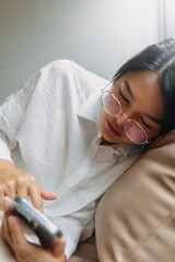 Obraz na płótnie Canvas Side view of asian Thai office woman wear glasses using mobile phone while leaning and resting on brown couch sofa, tired of work. 