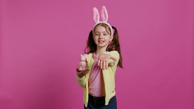 Lovely little girl with bunny ears sending air kisses in studio, presenting a stuffed pink rabbit toy for easter holiday. Smiling energetic preteen feeling positive and carefree. Camera B.