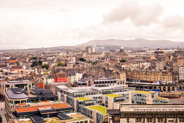 The streets and buildings of Edinburgh stretch out, forming a dynamic tapestry of urban life against the dramatic backdrop of Scotland's mountainous terrain. 
