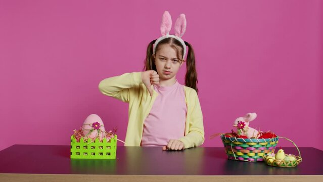 Displeased unhappy child giving thumbs down in studio, expressing her disapproval and disagreement with dislike symbol. Angry dissatisfied young girl showing negative bad gesture. Camera B.