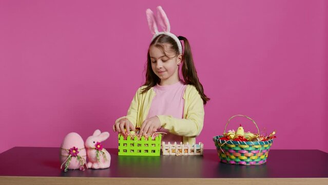 Joyful little girl showing her handcrafted festive basket on camera, creating a colorful handmade arrangement in time for easter holiday. Small toddler with bunny ears decorating eggs. Camera B.