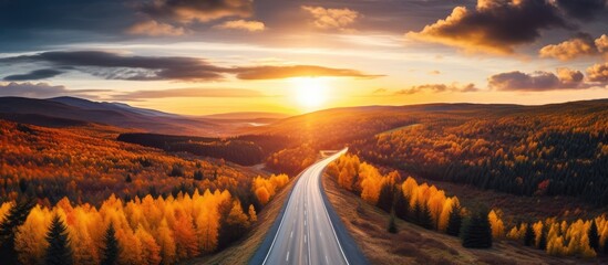 An aerial perspective of a road cutting through a vibrant forest during autumn, showcasing a stunning landscape under the warm hues of the setting sun.