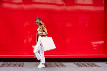 Stylish woman with shopping bags walks by the big red wall in the city