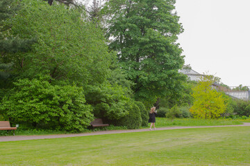 Young woman walking in green spring nature park. Distance view of girl in black dress and hat with trees on background