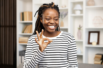Happy Woman Gesturing Ok Sign at Home. Afro American Female in striped shirt at home, expressing satisfaction and positive emotions in a cozy interior. - obrazy, fototapety, plakaty