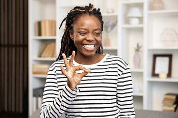 Happy Woman Gesturing Ok Sign at Home. Afro American Female in striped shirt at home, expressing satisfaction and positive emotions in a cozy interior.