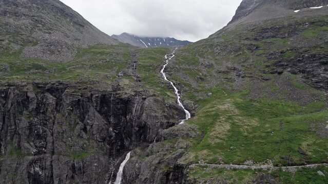 Wide aerial dolly shot, pushing in slowly on a long, thin, rushing river, winding down a steep, mossy hillside by Trollstigen, Norway. The steep hillside makes way for sheer cliffs in the foreground.