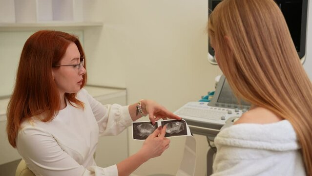 Tracking shot of professional female doctor checking results of ultrasound examination and explaining mage to young woman patient. Doctor explaining results of treatment by pointing on scan.