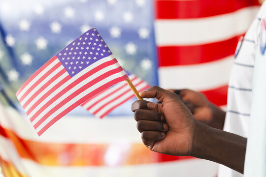 African American man's hand holding a small American flag, with a larger flag in the background