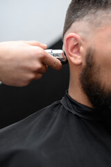 Barber shaves the temple with cordless trimmer during a short haircut on the sides of the head. - obrazy, fototapety, plakaty