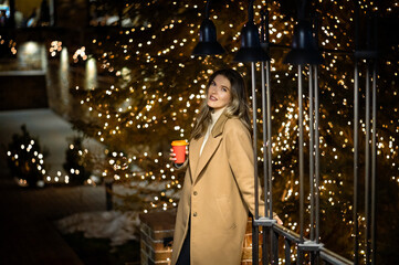 beautiful young girl in a beige coat with a paper glass of coffee against the backdrop of many...