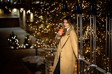 beautiful young girl in a beige coat with a paper glass of coffee against the backdrop of many...