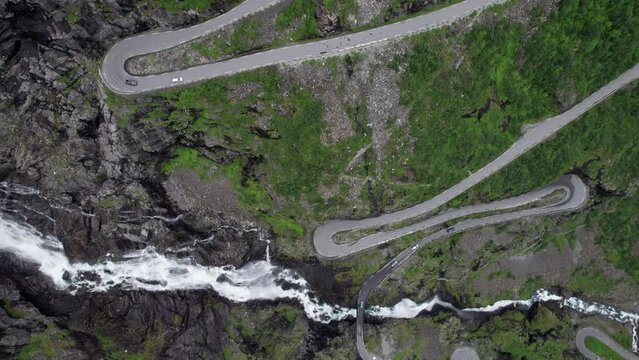 Bird's eye Aerial view showing the tight hairpin bends of the Trollstigen road in Norway. The Stigsfossen waterfall can be seen. Many cars drive on the road. Loose rocks and trees by the road.