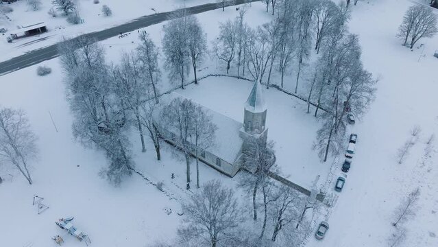 Snow-covered church in Renda village (Kuldiga parish, Latvia) with surrounding bare trees, aerial view, moving forward, tilt down