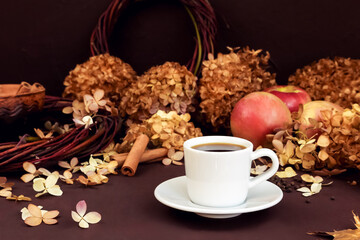 White coffee cup with espresso, cinnamon, red apples and dry hydrangea on dark brown background. autumn composition