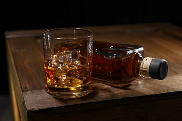 Whiskey with ice cubes in glass and bottle on wooden crate against black background, closeup