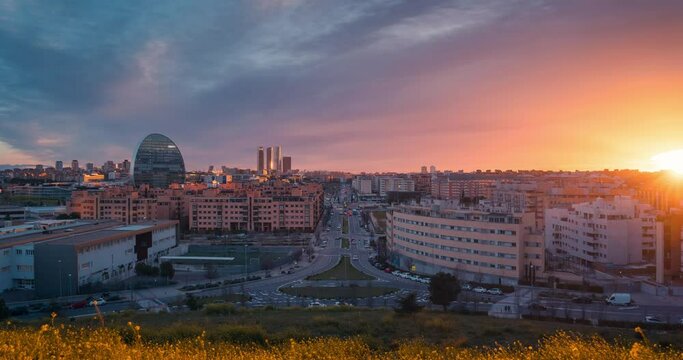 Timelapse skyline of modern european city Madrid from Las Tablas viewpoint during sunset with golden light and red clouds during blue hour zoom in day to night time lapse yellow flowers foreground