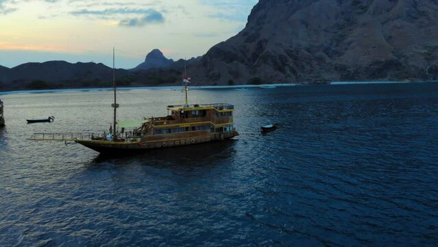 A cruise ship anchored off the coast of Padar Island, near Komodo in Indonesia, captured from a drone during the evening. In the background, a mountainous island adds to the picturesque scenery.