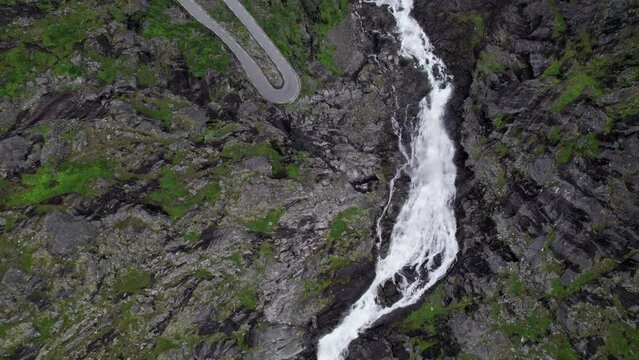 Top down aerial shot of the Trollfossen waterfall cascading down a cliff into the Tverelva river in Norway. Hairpin bends of the serpentine Trollstigen mountain road can be seen next to the waterfall.