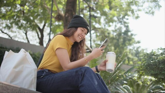 Gen Z eco go green woman asia sit happy relax at tree public park city life enjoy typing mobile phone drink coffee bottle use reuse tote bag care save the earth planet net zero waste clean future hope