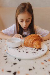 Cheerful preschool girl having breakfast in the cafe. The teen is eating a croissant. The concept of tourism with a child and healthy eating