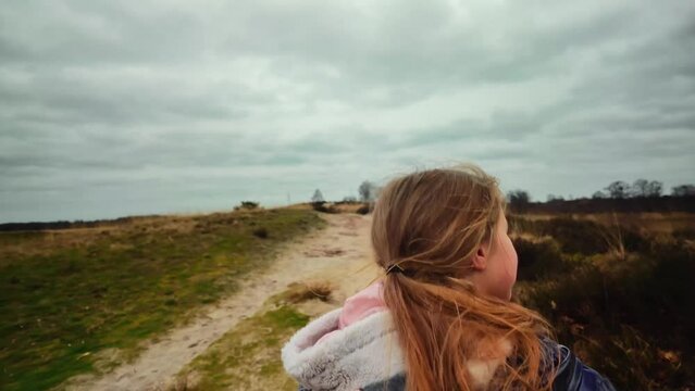 Blond girl with ponytale walking on dune hill with sand on cloudy windy day