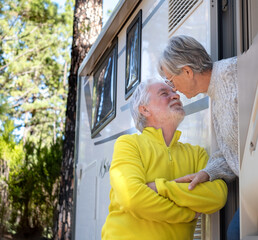 Smiling senior couple outside their camper van motor home exchange kisses enjoying retirement lifestyle. Elderly people and freedom vacation travel in the forest.