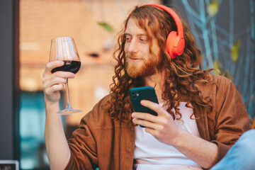 A red hair man with music headphones holding a glass of wine and a cell phone