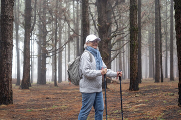 Active mature woman with backpack walking in mountain forest on a foggy day with the help of poles enjoying nature, freedom and free time. Forest background with bare trees
