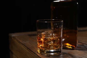 Whiskey with ice cubes in glass and bottle on wooden crate against black background, closeup. Space for text