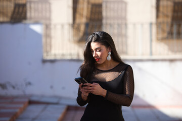 Young, beautiful brunette woman is dressed elegantly in a black dress with transparencies and fancy earrings. The girl is walking and consulting her phone. The woman is wearing make-up with red lips.