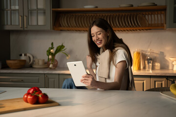 A young woman sits at a kitchen counter bathed in warm sunlight, visibly elated as she celebrates a win on her tablet device. Fresh vegetables, including bright red tomatoes, are nearby, suggesting a