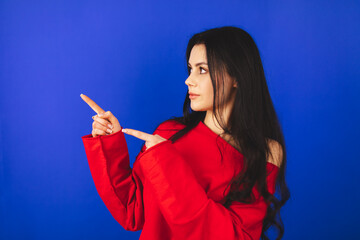 Fototapeta premium Look, advertise here! Dark haired pretty woman with appealing smile gives positive attitude towards good deal, expresses her recommendation while pointing on left side isolated on blue background.