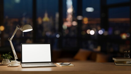 Office at night, a laptop computer mockup, a table lamp, and accessories on a wooden desk.