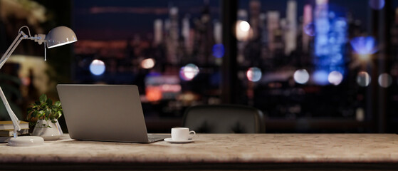 A modern office at night with a city view, featuring a laptop and accessories on a desk.