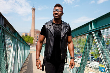 Portrait of smiling young stylish african american man wears eyeglasses and leather sleeveless jacket, standing on bridge looking at camera.