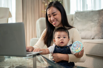 A positive Asian mom is playing with and taking care of her little son while working on her laptop.