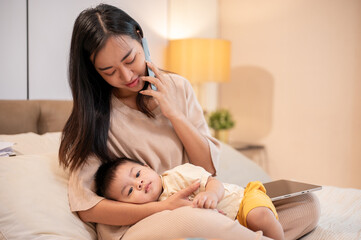 Obraz na płótnie Canvas A caring Asian mom is putting her little son to sleep in her arms while she is talking on the phone.