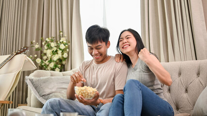 A cheerful Asian couple is watching an exciting sports match or game show together at home. - obrazy, fototapety, plakaty