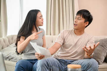 A resentful, angry Asian wife and husband are arguing about family household invoices on sofa.
