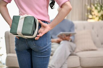 A close-up, back view image of a lovely wife hiding a surprise gift box behind her from her husband.