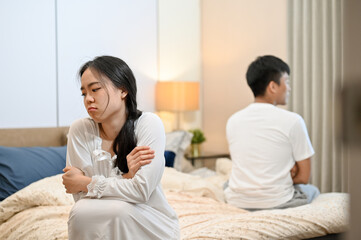 An unhappy, sad young Asian couple in pajamas is sitting separately on the bed after their argument.