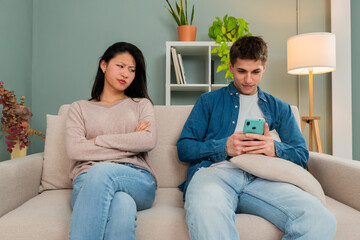 Young couple sitting on the couch, he is using the mobile phone and she is peeking him with upset expression. Boyfriend and gilrdfriend with communication problems arguing in home interior - obrazy, fototapety, plakaty