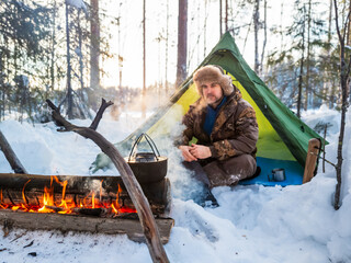 Man Enjoying a Warm Campfire Outside His Tent in a Snowy Forest at Dusk. Dressed for winter weather, he rests beside his tent pitched amidst a snowy landscape