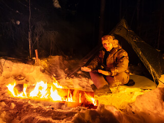Man Sitting Next to Long Fire in Snow.  A man sits next to a crackling fire in a snowy setting, warming himself in the cold winter weather. The snow-capped landscape contrasts