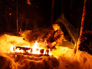 Man Sitting Next to Long Fire in Snow.  A man sits next to a crackling fire in a snowy setting,...