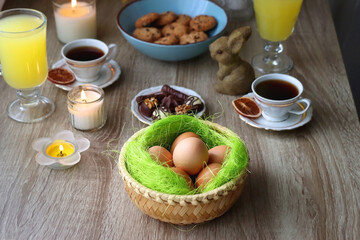 Easter eggs in the basket, bowl of cookies, chocolate pralines, Easter bunny figurine, cups of tea, glasses of juice, flowers and lit candles on the table. Selective focus.